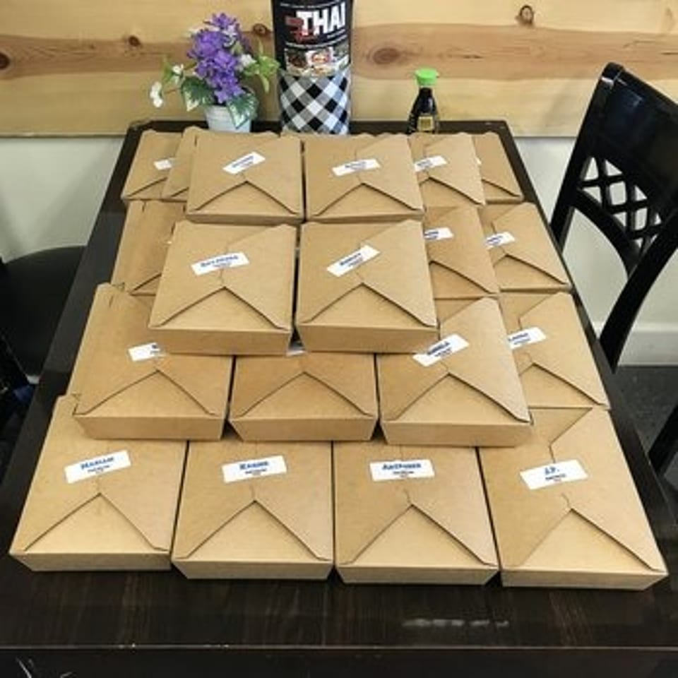 Table covered with labeled brown takeout boxes, alongside a Thai drink cup, flowers, and dining chairs.