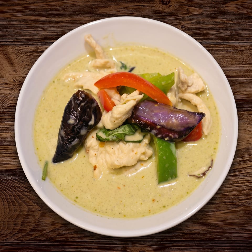 Bowl of creamy green curry with chicken, eggplant, bell peppers, and basil on a wooden table.