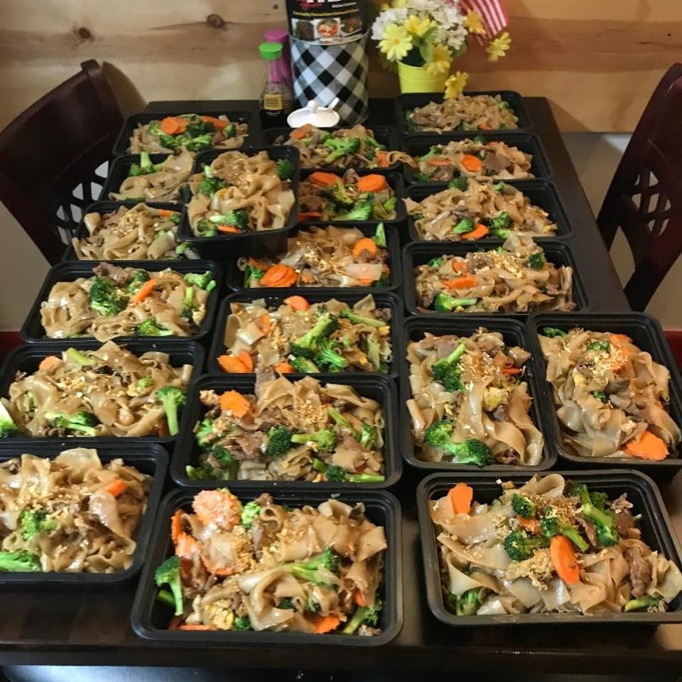 Numerous black takeout containers hold Thai stir-fried noodles with vegetables, arranged on a restaurant table beside flowers and American flags.