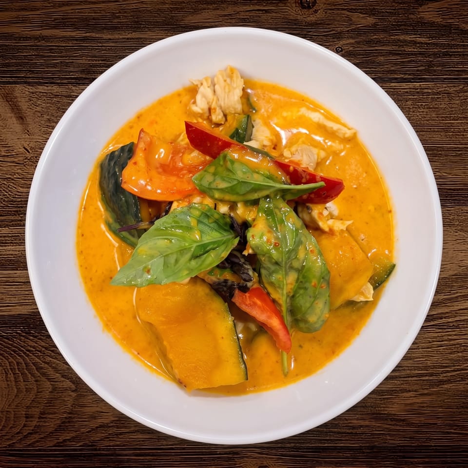 Thai red curry with chicken, vegetables, red peppers, and fresh basil in a white bowl on a wooden table.