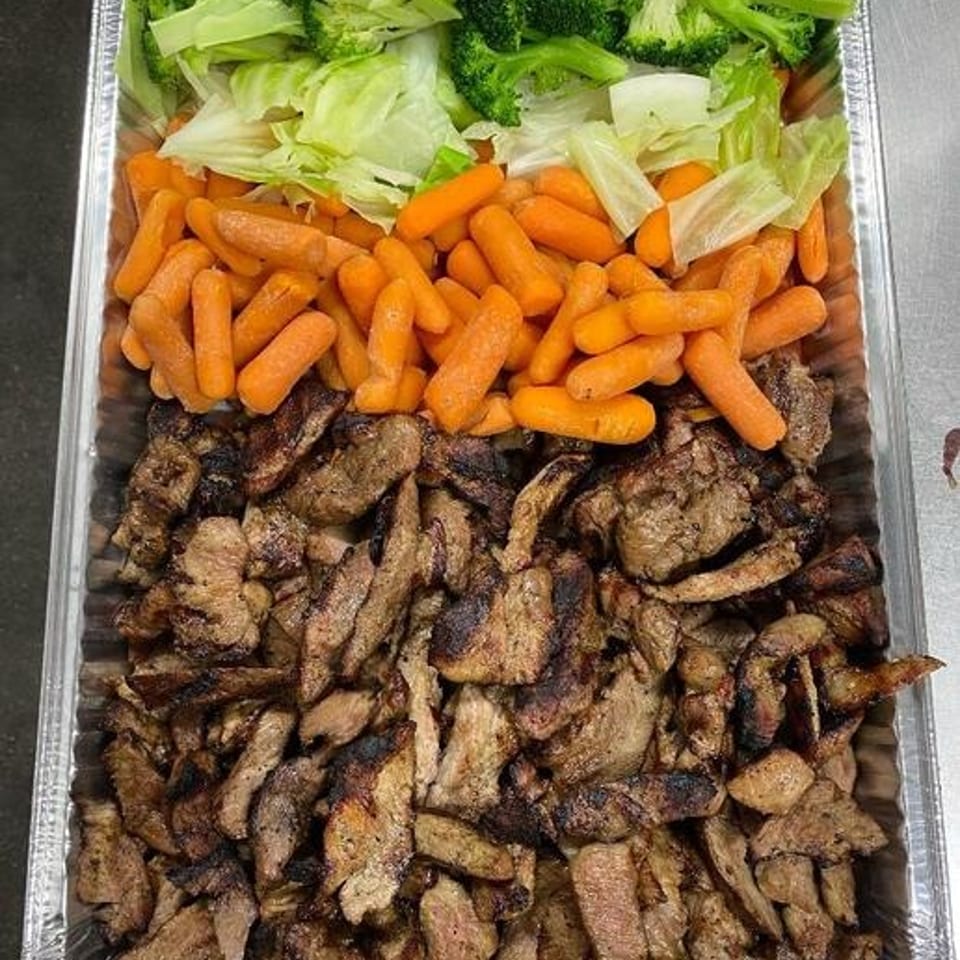 Aluminum catering tray filled with grilled sliced meat, baby carrots, iceberg lettuce, and broccoli.