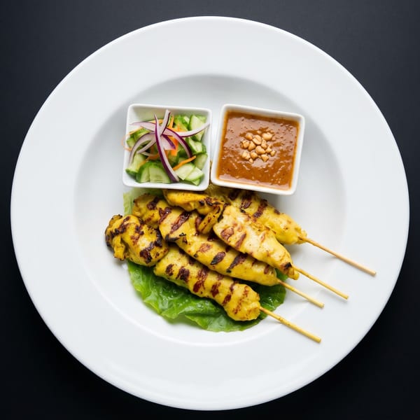 Grilled chicken satay skewers with peanut sauce and cucumber salad on a white plate.